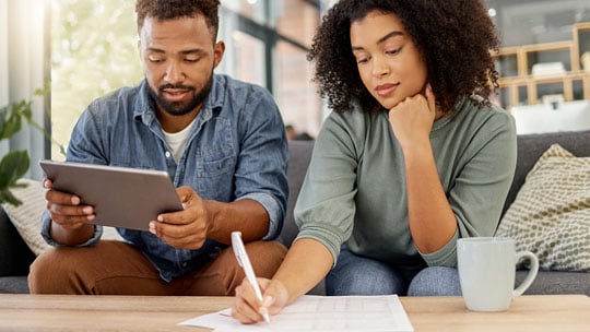 Man sitting on couch with table next to a woman taking notes with pen and paper.